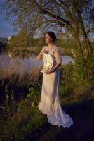 un' donna nel un' bianca Vintage ▾ vestito su il riva di un' lago a tramonto detiene un' Luna sagomato specchio foto