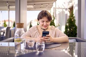 più dimensione donna nel pesca fuzz vestito sceglie un' piatto nel un' ristorante attraverso qr codice nel smartphone, acqua con Limone e bicchieri nel davanti di sua foto
