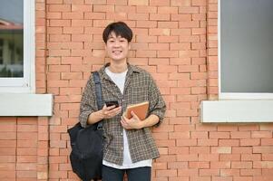 sorridente asiatico maschio Università alunno con un' zaino sta contro un' mattone parete di un' città universitaria costruzione. foto