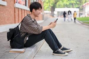 un' positivo asiatico maschio Università alunno si siede su il strada vicino il città universitaria edificio utilizzando il suo Telefono. foto