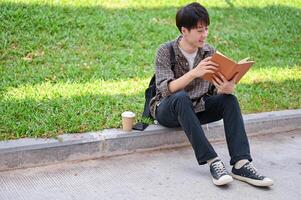 un' positivo asiatico maschio Università alunno si siede su il strada nel il città universitaria parco lettura un' libro. foto