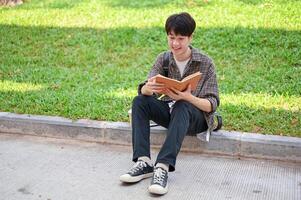 un' positivo asiatico maschio Università alunno si siede su il strada nel il città universitaria parco lettura un' libro. foto