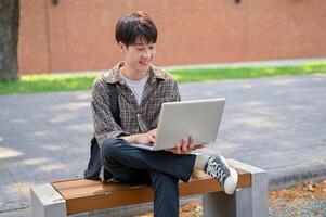 un' giovane asiatico maschio Università alunno utilizzando il suo il computer portatile computer su un' panchina nel il città universitaria parco. foto