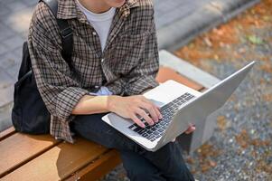 un' ritagliata Immagine di un' maschio Università alunno utilizzando il suo il computer portatile computer su un' panchina nel il città universitaria parco. foto