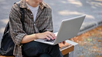 un' ritagliata Immagine di un' maschio Università alunno utilizzando il suo il computer portatile computer su un' panchina nel il città universitaria parco. foto