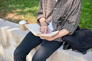 un' ritagliata Immagine di un' maschio Università alunno si siede su un' pietra panchina nel un' parco, lettura un' libro all'aperto. foto