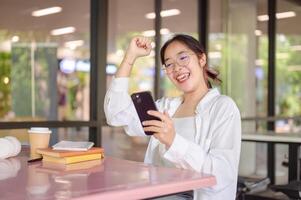 un eccitato asiatico femmina Università alunno celebra bene notizia mentre guardare a sua Telefono a un' tavolo. foto