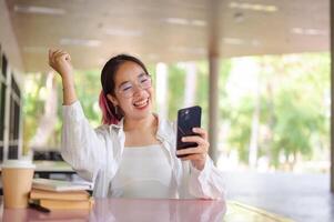 un eccitato asiatico femmina Università alunno celebra bene notizia mentre guardare a sua Telefono a un' tavolo. foto