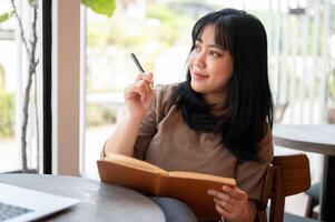 un' positivo asiatico femmina è conservazione sua diario o listato sua idee nel un' libro mentre seduta nel un' bar. foto