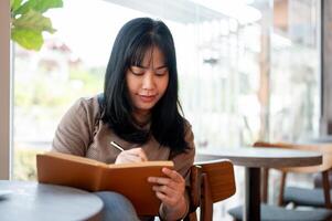 un' positivo asiatico femmina è conservazione sua diario o listato sua idee nel un' libro mentre seduta nel un' bar. foto