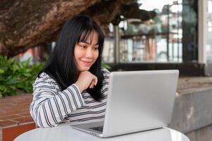 un' sorridente e positivo asiatico donna è Lavorando remoto all'aperto nel un' città parco, utilizzando sua il computer portatile. foto