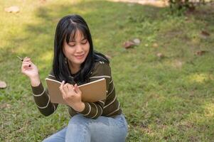 un' positivo giovane asiatico donna seduta su il erba nel un' parco con sua diario, conservazione sua diario. foto