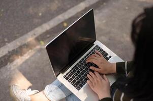 un' avvicinamento Immagine di un asiatico donna Lavorando remoto nel un' parco, utilizzando sua il computer portatile computer su un' panca. foto