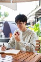 un' bello asiatico uomo è mangiare un Mela mentre scrittura giù il suo idee nel un' libro a un' bar. foto