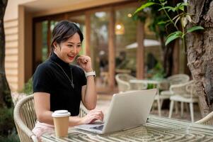 un' allegro donna si siede a un' tavolo, guardare a sua il computer portatile con un' contento viso, festeggiare bene notizia. foto