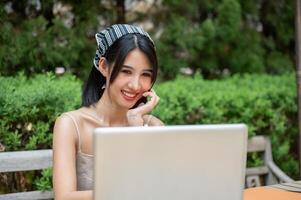 un attraente, positivo asiatico donna Lavorando su sua il computer portatile computer a un' tavolo nel un' verde giardino. foto