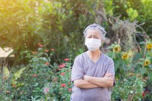 un' ritratto di un anziano donna indossare un' viso maschera e braccia attraversato mentre in piedi nel un' giardino. maschera per proteggere virus, coronavirus, polline grani. concetto di vecchio persone e assistenza sanitaria. foto