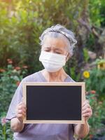 ritratto di anziano asiatico donna Tenere un' lavagna e indossare un' viso maschera mentre in piedi nel il giardino. maschera per protezione virus, covid19, coronavirus. concetto di anziano persone e assistenza sanitaria foto