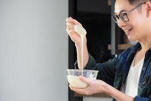 un' giovane uomo indossare casuale capi di abbigliamento uso un' frusta miscelazione ingredienti nel un' ciotola mentre in piedi nel il cucina. cucina utensili e cucinando concetto foto