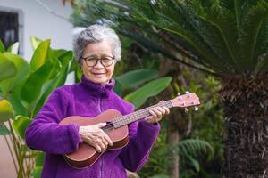 ritratto di un anziano donna giocando il ukulele mentre in piedi nel il giardino. rilassante di cantando e giocare piccolo chitarra contento e godere vita dopo andare in pensione. concetto di anziano persone e assistenza sanitaria foto