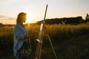 silhouette di un' bionda ragazza artista. signora vernici un' pittura su il tela con il Aiuto di vernici. un' di legno cavalletto mantiene il immagine. estate è un' soleggiato giorno, tramonto. foto