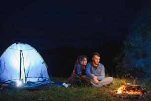 notte campeggio nel il montagne. contento coppia i viaggiatori seduta insieme accanto fuoco di bivacco e raggiante turista tenda. su sfondo grande masso, foresta e notte cielo. foto