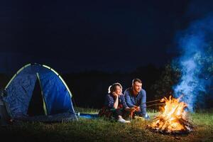 campeggio notte nel montagne. turista coppia seduta nel davanti di illuminato tenda illuminato di ardente falò. turismo e all'aperto attività concetto. foto