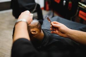lato Visualizza di grave uomo con elegante moderno taglio di capelli guardare inoltrare nel barbiere negozio. mano di barbiere conservazione dritto rasoio e taglio di moda strisce su testa di cliente. foto