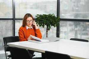 contento sorridente attività commerciale donna a opera parlando su Telefono, seduta a sua Lavorando posto nel ufficio, copia spazio foto