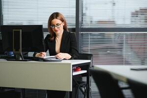 bello, simpatico, carino, Perfetto donna seduta a sua scrivania su pelle sedia nel opera stazione, indossare occhiali, vestito formale, avendo il computer portatile e taccuino su il tavolo foto