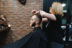 servendo cliente nel barbiere. professionale barbiere ragazza, femmina parrucchiere fabbricazione moderno taglio di capelli per un' uomo seduta nel barbiere negozio sedia. messa a fuoco su un' ragazza. parrucchiere, rasatura, Rifinitura, toelettatura. foto