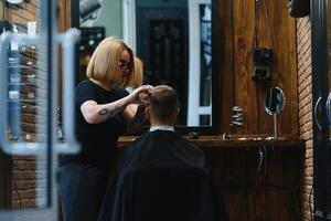 elegante uomo seduta barbiere negozio parrucchiere parrucchiere donna taglio il suo capelli ritratto bello contento giovane barbuto caucasico tipo ottenere di moda taglio di capelli attraente barbiere ragazza Lavorando servendo cliente. foto