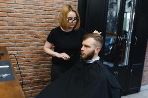 elegante uomo seduta barbiere negozio parrucchiere parrucchiere donna taglio il suo capelli ritratto bello contento giovane barbuto caucasico tipo ottenere di moda taglio di capelli attraente barbiere ragazza Lavorando servendo cliente foto