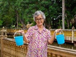 ritratto di un anziano asiatico donna Tenere un' blu plastica benna, sorridente e guardare a il telecamera mentre in piedi lato il stagno. concetto di anziano persone e assistenza sanitaria foto