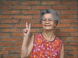 un anziano asiatico donna con corto grigio capelli, sorridente, mostrando vittoria cartello e guardare a il telecamera mentre in piedi con mattone parete sfondo. concetto di anziano persone e assistenza sanitaria foto