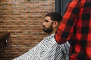 fabbricazione taglio di capelli Guarda Perfetto. giovane barbuto uomo ottenere taglio di capelli di parrucchiere mentre seduta nel sedia a barbiere. foto