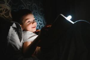 lettura libro e utilizzando torcia elettrica. giovane ragazzo nel casuale Abiti dire bugie giù vicino tenda a sera tempo foto