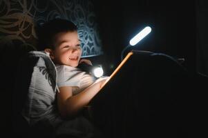 bambino lettura libro nel letto. bambini leggere a notte. poco ragazzo con Fata racconto libri nel Camera da letto . formazione scolastica per giovane bambini. di andare a dormire storia nel il sera. carino ragazzo sotto coperta nel buio camera con lampada foto