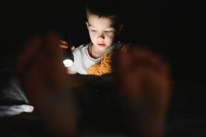 bambino lettura libro nel letto. bambini leggere a notte. poco ragazzo con Fata racconto libri nel Camera da letto . formazione scolastica per giovane bambini. di andare a dormire storia nel il sera. carino ragazzo sotto coperta nel buio camera con lampada foto