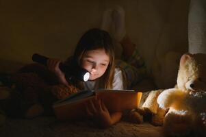 contento bambino ragazza ridendo e lettura libro nel buio nel un' tenda a casa foto