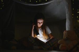 poco ragazza siamo lettura un' libro con torce elettriche nel tenda. contento ragazza giocando a casa. divertente bello ragazzo avendo divertimento nel camera. foto