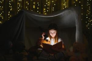 contento bambino ragazza ridendo e lettura libro nel buio nel un' tenda a casa foto