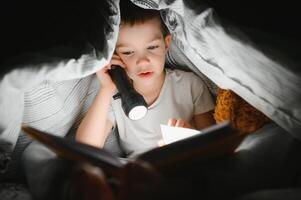 lettura libro e utilizzando torcia elettrica. giovane ragazzo nel casuale Abiti dire bugie giù vicino tenda a sera tempo foto