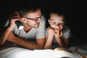 padre e figlio con torcia elettrica lettura libro sotto coperta a casa. foto