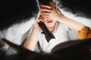 bambino lettura libro nel letto. bambini leggere a notte. poco ragazzo con Fata racconto libri nel Camera da letto . formazione scolastica per giovane bambini. di andare a dormire storia nel il sera. carino ragazzo sotto coperta nel buio camera con lampada foto