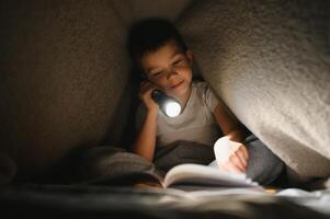 bambino lettura libro nel letto. bambini leggere a notte. poco ragazzo con Fata racconto libri nel Camera da letto . formazione scolastica per giovane bambini. di andare a dormire storia nel il sera. carino ragazzo sotto coperta nel buio camera con lampada foto