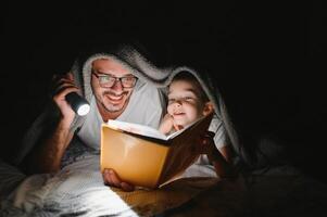 padre e figlio con torcia elettrica lettura libro sotto coperta a casa. foto