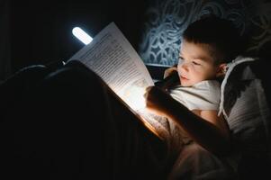 bambino lettura libro nel letto. bambini leggere a notte. poco ragazzo con Fata racconto libri nel Camera da letto . formazione scolastica per giovane bambini. di andare a dormire storia nel il sera. carino ragazzo sotto coperta nel buio camera con lampada foto