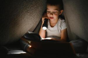 ritratto di carino poco ragazzo lettura nel letto con torcia elettrica nel buio camera, godendo fiabe foto