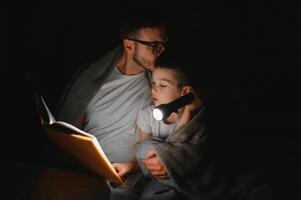 padre e figlio con torcia elettrica lettura libro sotto coperta a casa. foto